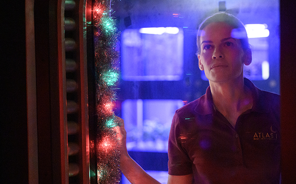A woman in a maroon polo shirt looks through a window adorned with glowing red and green tinsel lights.