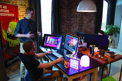 A man stands gesturing toward a seated man working on two computer monitors in an office.