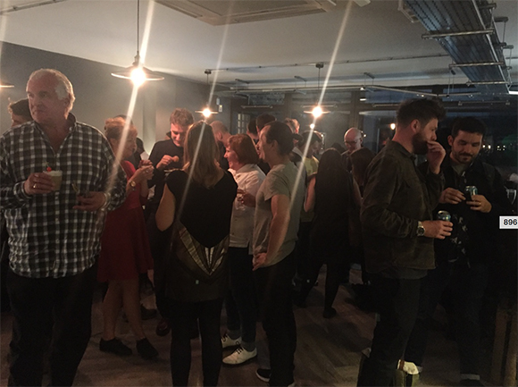 A crowd of people standing and talking in a dimly lit room with hanging lights.