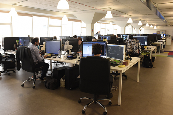 People sitting at desks with multiple computer monitors in a large, brightly lit open-plan office.