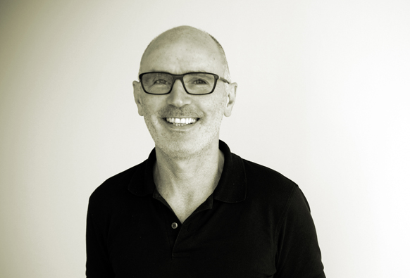 A smiling man with glasses and a black polo shirt against a light background.
