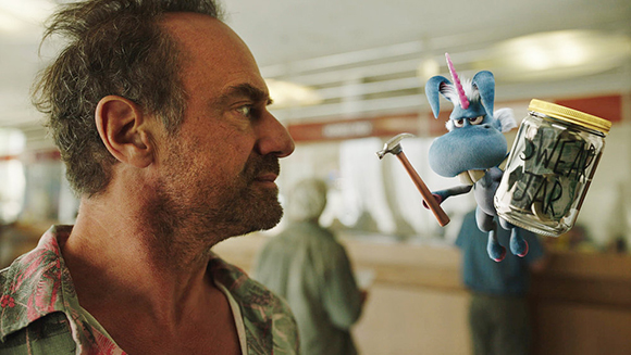 A man looks intensely at a small blue unicorn figurine holding a hammer and a jar labeled 'SWEAR JAR'.