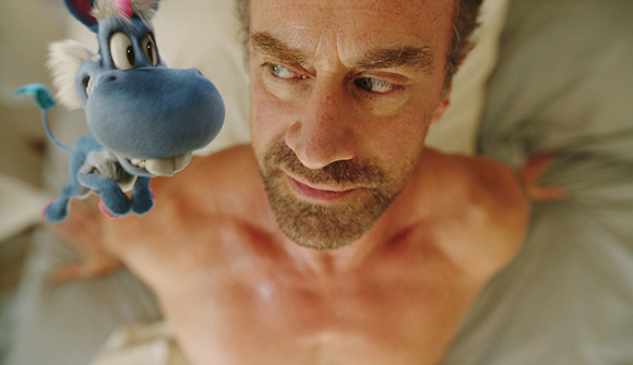 A man lies in bed looking up at a small, blue, animated donkey puppet positioned near his face.
