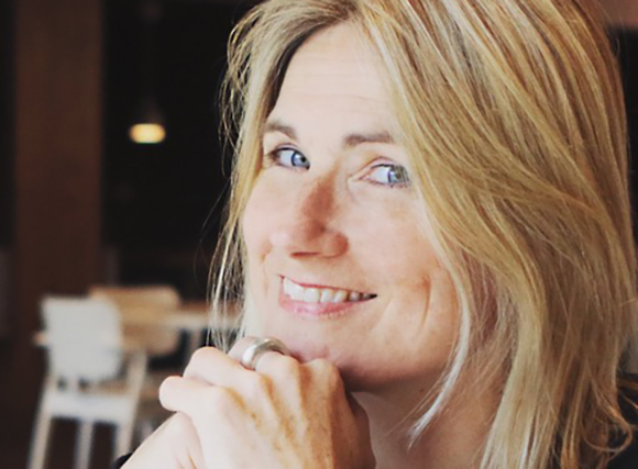 A smiling blonde woman rests her chin on her hands while looking toward the camera.