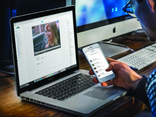 A person holds a smartphone while looking at a laptop screen displaying a video player.