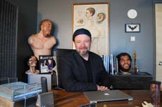 A man in a dark jacket and beanie sits at a desk with a laptop, surrounded by busts and artwork.