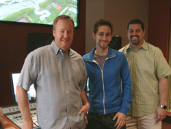 Three men smiling and standing together in a studio with a mixing console and monitor in the background.