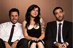 A woman with dark hair and tattoos sits between two men in formal attire.