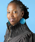 A woman with dark braided hair and large circular blue earrings wearing a grey high-collared jacket.
