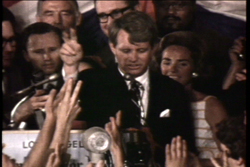 A man in a dark suit speaks at a podium while a crowd of people watches and raises their hands.