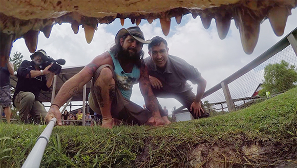 Two men smile while crouching under the open jaws of an alligator on a grassy area.