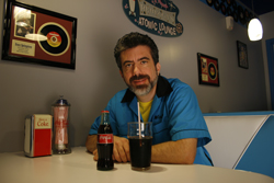 Man in a blue shirt sitting at a diner table with a glass of cola and a glass bottle.