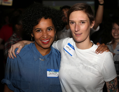 Two women smiling, one in a blue denim shirt and one in a white t-shirt, both wearing name tags.