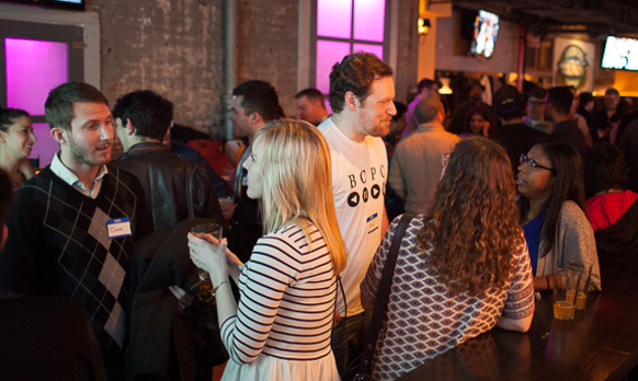 A diverse group of people talking and holding drinks in a crowded, dimly lit indoor venue.