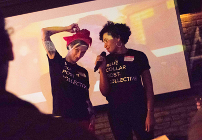 Two people wearing black Blue Collar Post Collective t-shirts stand in front of a large projection screen.