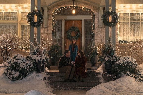 A man and woman sit on stone steps in front of a house decorated with wreaths and string lights in the snow.