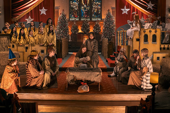 Children in costumes performing a nativity play on a stage with Christmas trees and stained glass background.