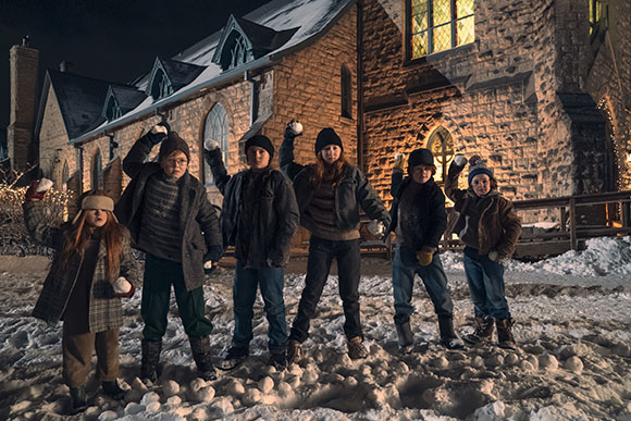 Six children in winter clothing stand in the snow, holding snowballs, in front of a large stone building at night.