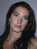 A woman with long dark hair and silver hoop earrings looks toward the camera against a plain background.