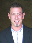 A smiling man with short dark hair wearing a white patterned shirt and a black blazer.