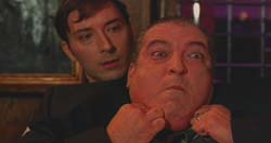 A man in a dark suit pulls the cheeks of another man, making his face bulge.