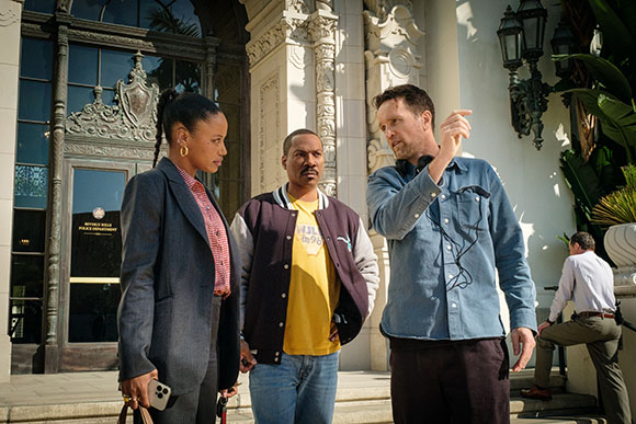 A woman in a blazer, a man in a varsity jacket, and a man in a denim shirt talk outside a building.