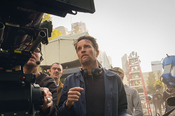 A man in a blue jacket stands on a film set looking toward a camera crew under a bright sky.