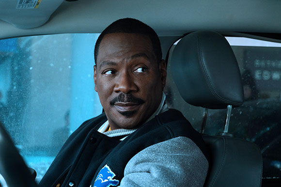 A man with a mustache sits in a car seat, looking back over his shoulder with a slight smile.