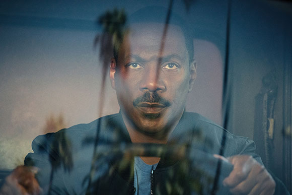 A man with a mustache looks forward through a window reflecting palm trees.