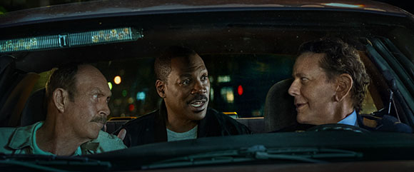 Three men sit inside a car at night, looking at each other with various expressions.