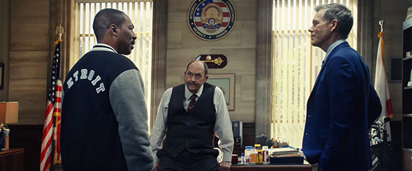 Three men in an office: one in a varsity jacket, one in a vest, and one in a blue suit.