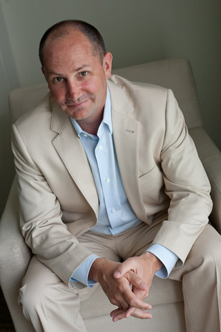 A man wearing a beige suit and light blue shirt sits in a cream-colored armchair.