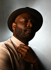 A man with a beard and hat poses against a smoky, backlit background.