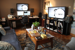 A living room featuring a desk with dual monitors, a large television, and a coffee table with books.