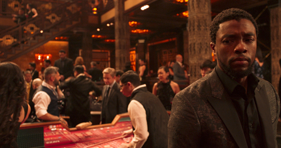 A man in a dark patterned suit looks forward in a crowded, dimly lit upscale lounge or casino.
