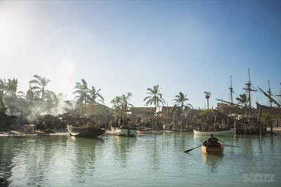 Small boats float in a calm tropical bay near a village with palm trees under a bright sky.