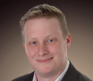 A smiling man with light hair and blue eyes wearing a dark suit jacket and light shirt.