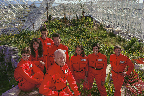 Nine people in matching red jumpsuits pose together amidst lush greenery inside a large geodesic dome.