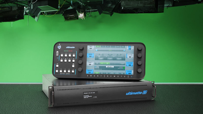 A black Ultimatte device with a touchscreen display and buttons sits on top of a black rackmount unit. A green screen is in t