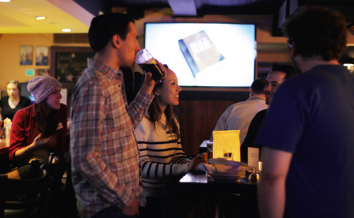 A group of people socializing in a bar, with a man drinking from a glass in the foreground.