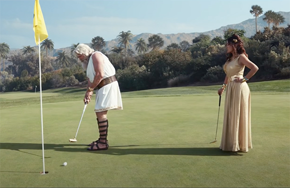 A man in a white tunic and sandals prepares to putt on a green, while a woman in a gold dress watches.