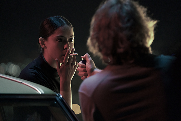 A woman in dark clothing lights a cigarette from a lighter held by a person in the foreground at night.