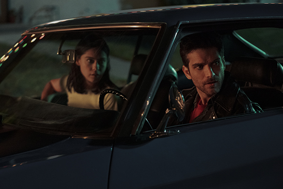 A man and a woman sit inside a dark car at night, looking ahead with serious expressions.