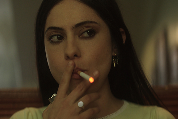 A woman with dark hair looks to the side while holding a lit cigarette to her lips.