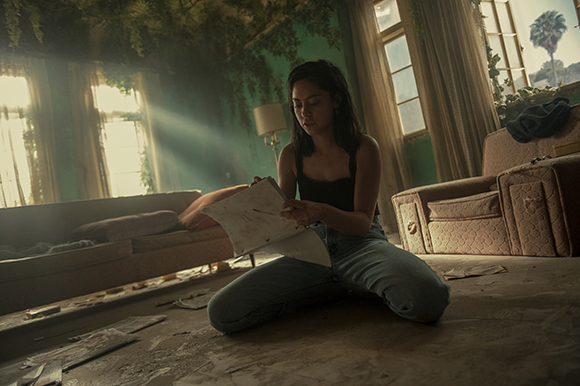 A woman sits cross-legged on a dusty floor, writing in a notebook in a room with overgrown plants.