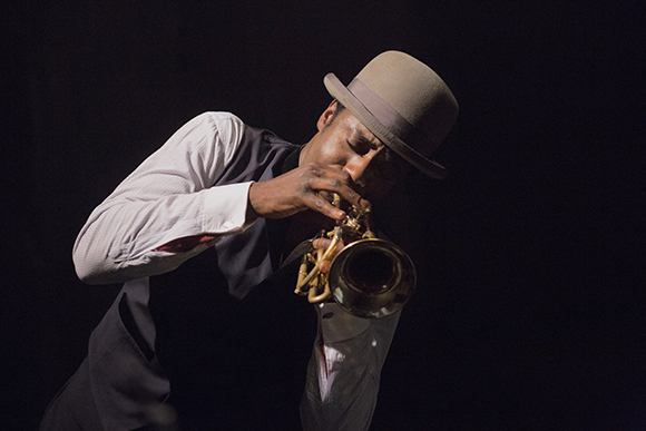 A man wearing a fedora and vest plays a brass instrument against a dark background.