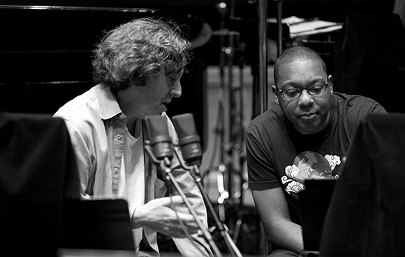A man with curly hair sings into a microphone while another man watches closely in a studio.