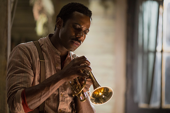 A man in a striped shirt and suspenders playing a brass trumpet in a dimly lit room.