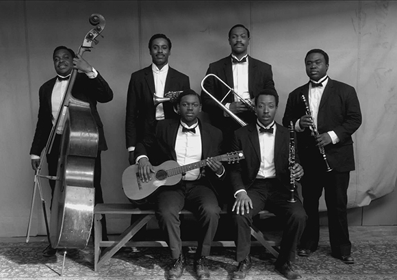 Six Black musicians in tuxedos pose with a double bass, trumpet, trombone, guitar, clarinet, and saxophone.