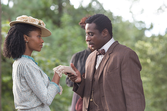A man in a brown suit holds the hand of a woman in a light blue dress and floral hat.